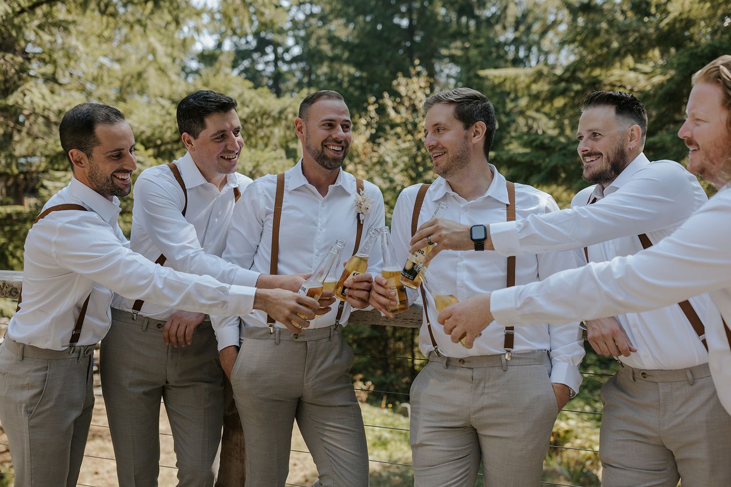 Groomsmen in light khaki suits toast and celebrate together outdoors.