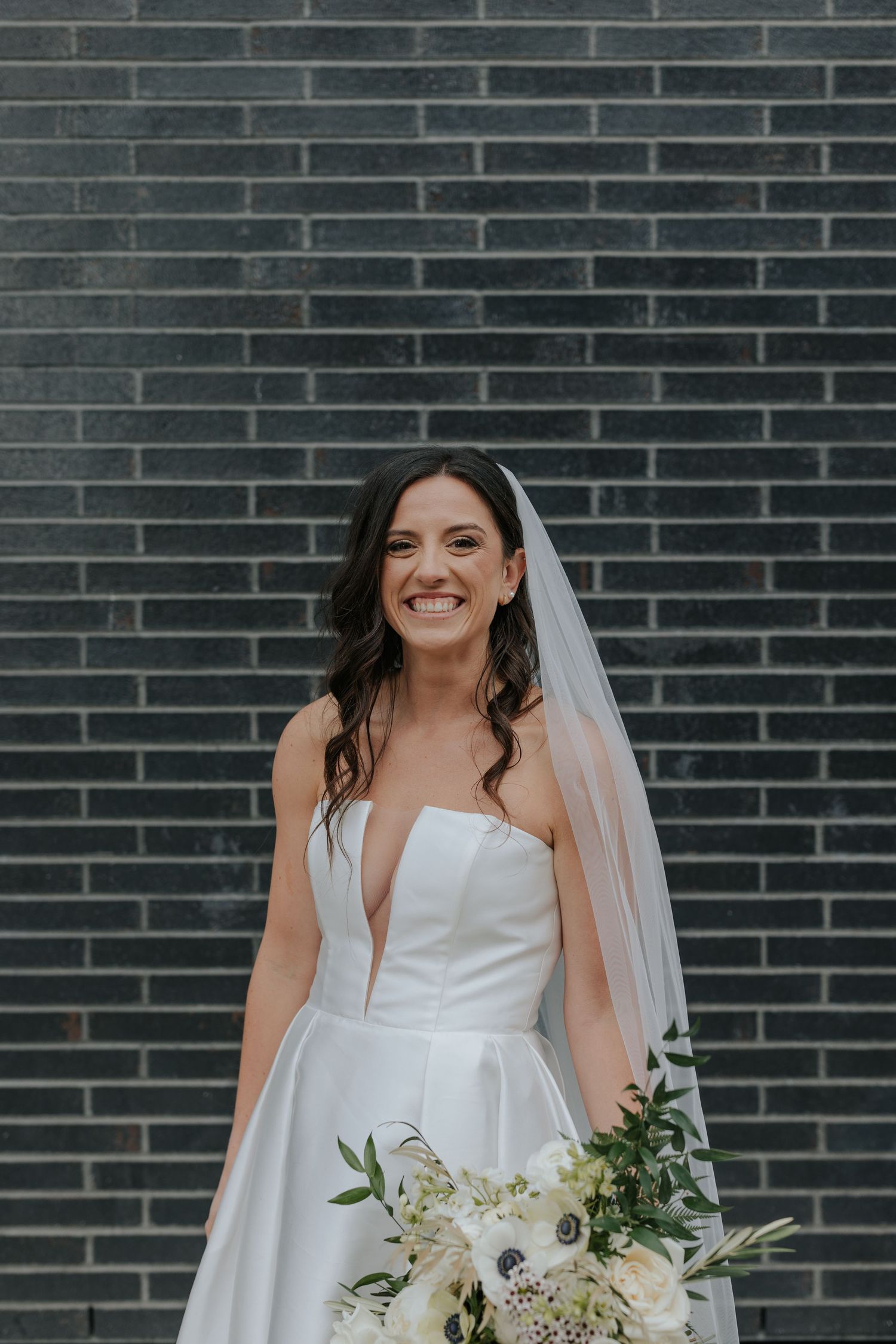 A bride in an elegant white wedding gown holds a beautiful bouquet against a dark brick wall backdrop.