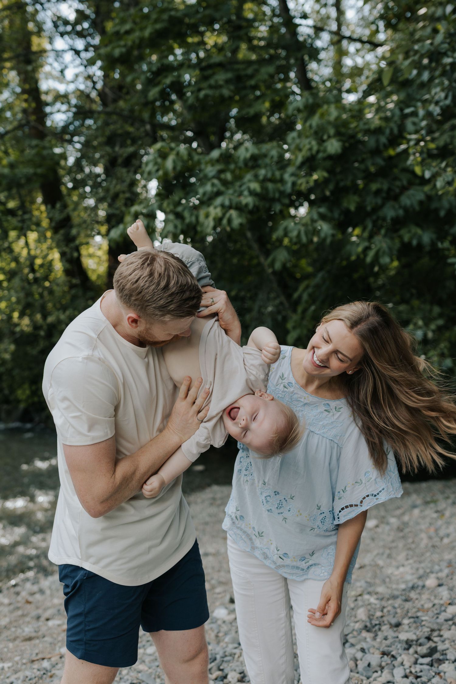 Happy young family with baby enjoying outdoor time near a rocky riverbank surrounded by lush green trees.