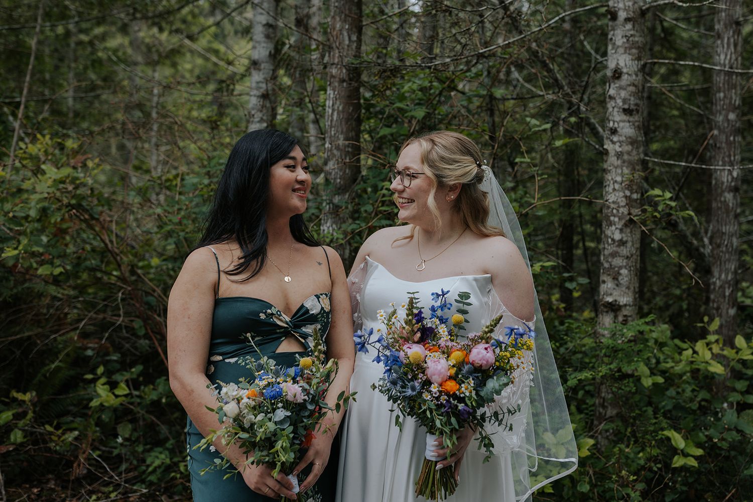 Wedding party members share laughs while holding colorful bouquets in a forest setting.