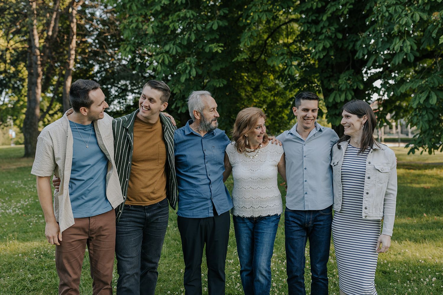 A multi-generational family group poses together outdoors in casual clothing against a green background.