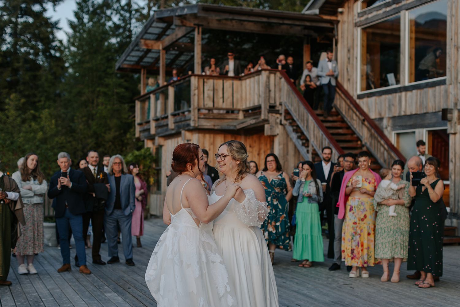 Wedding ceremony attendees stand on a wooden deck surrounded by mountains and evergreen trees.