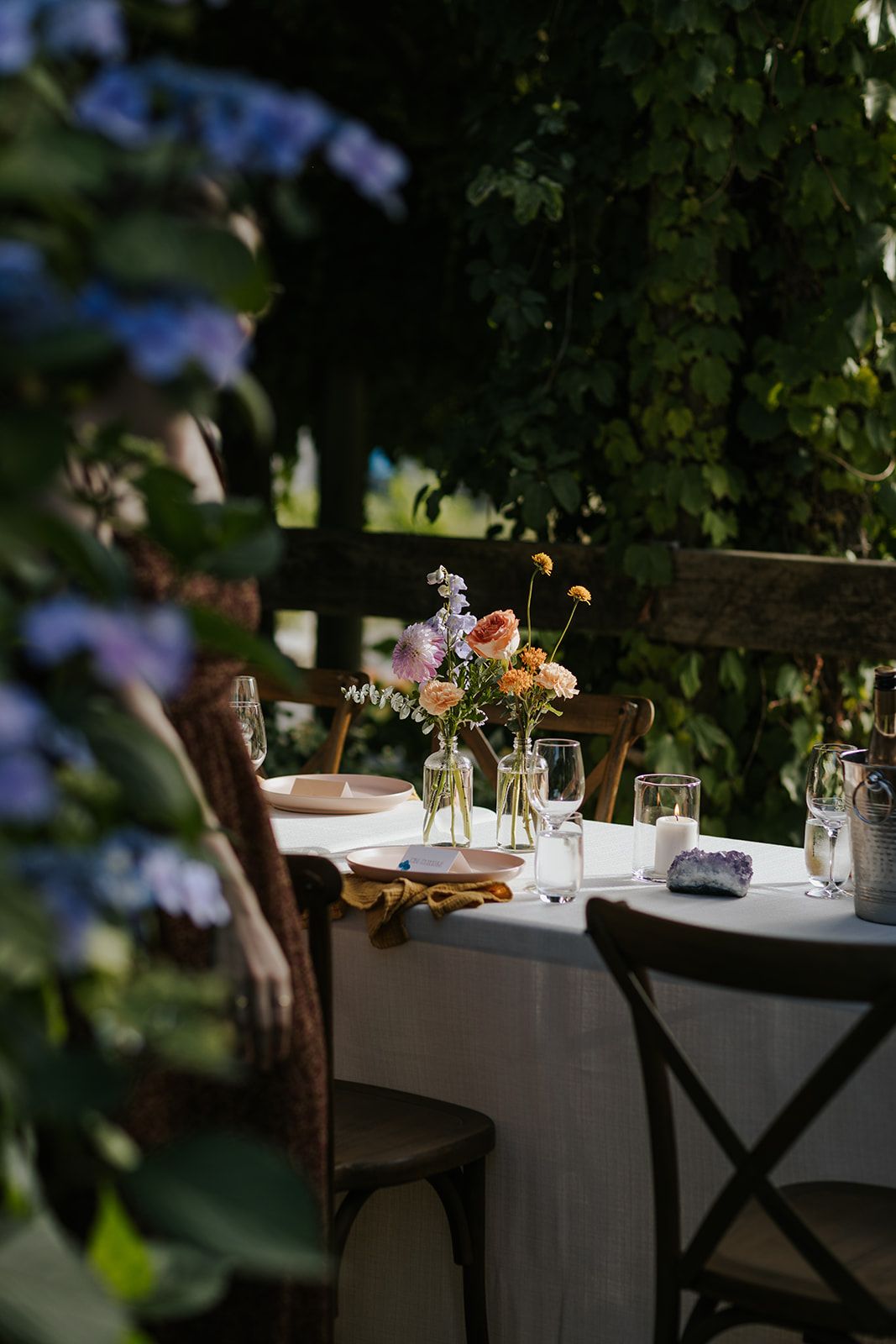 Outdoor dining tables set with white linens and floral centerpieces under garden trees.
