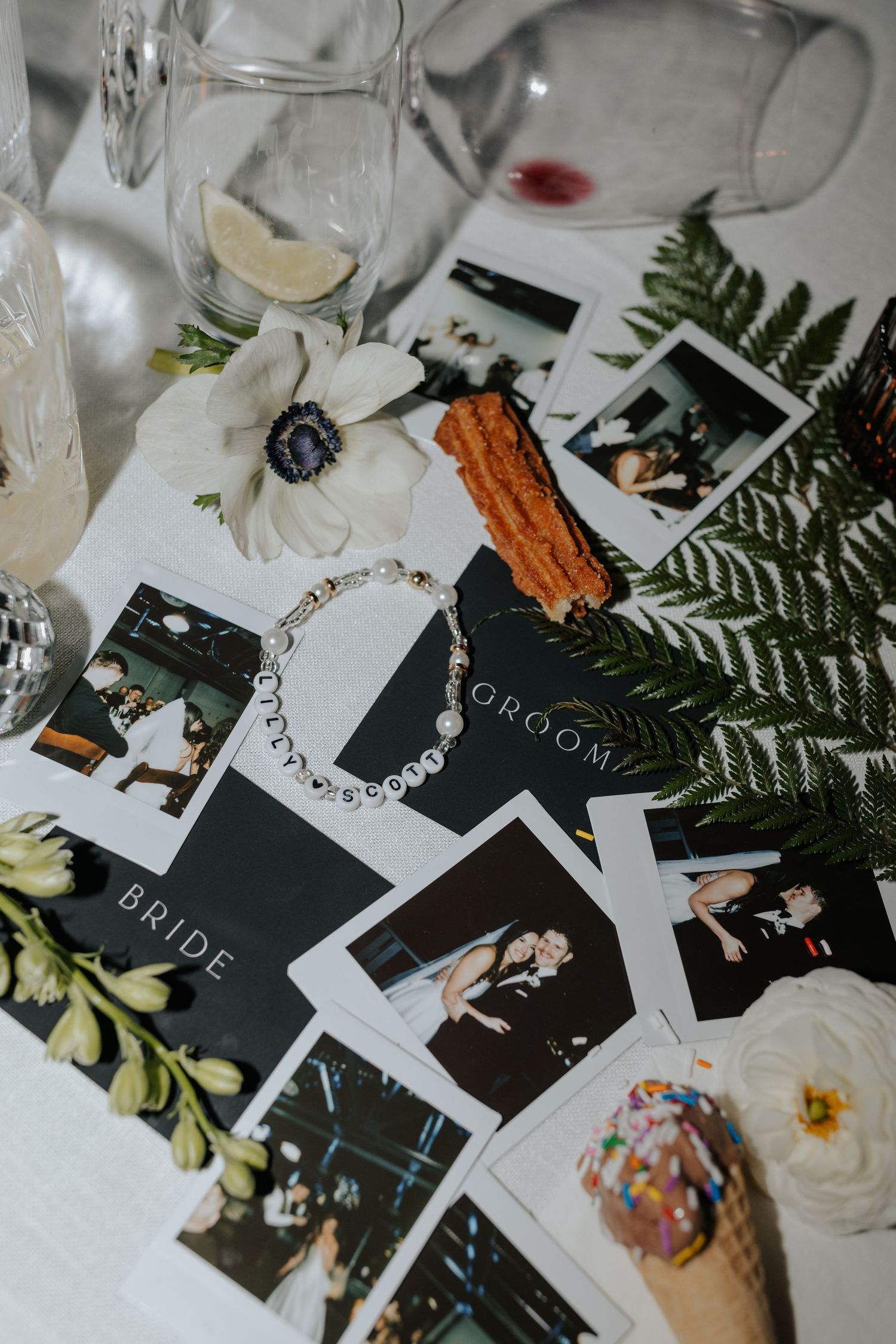 Wedding reception table details with scattered photos, ferns, and white floral decorations on a light surface.