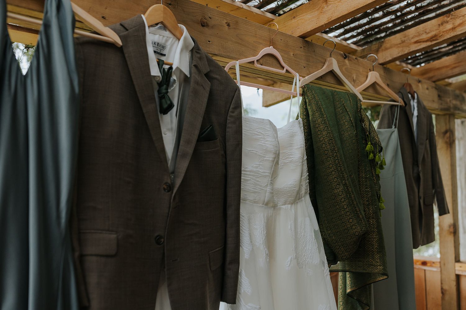 Wedding attire including suits and dresses displayed on hangers against wooden backdrop.