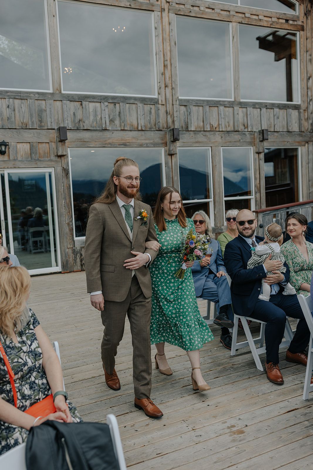 Couple walks down aisle at mountain venue while guests look on from white chairs.