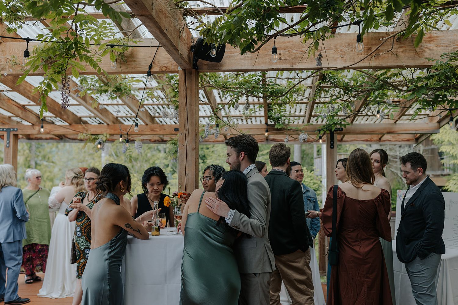 Wedding guests mingling under a vine-covered pergola at an outdoor reception.