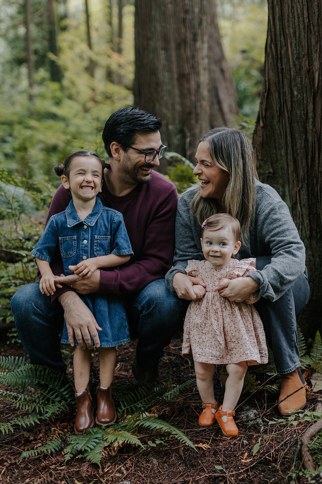 Family sits together on forest floor surrounded by ferns and tall trees.