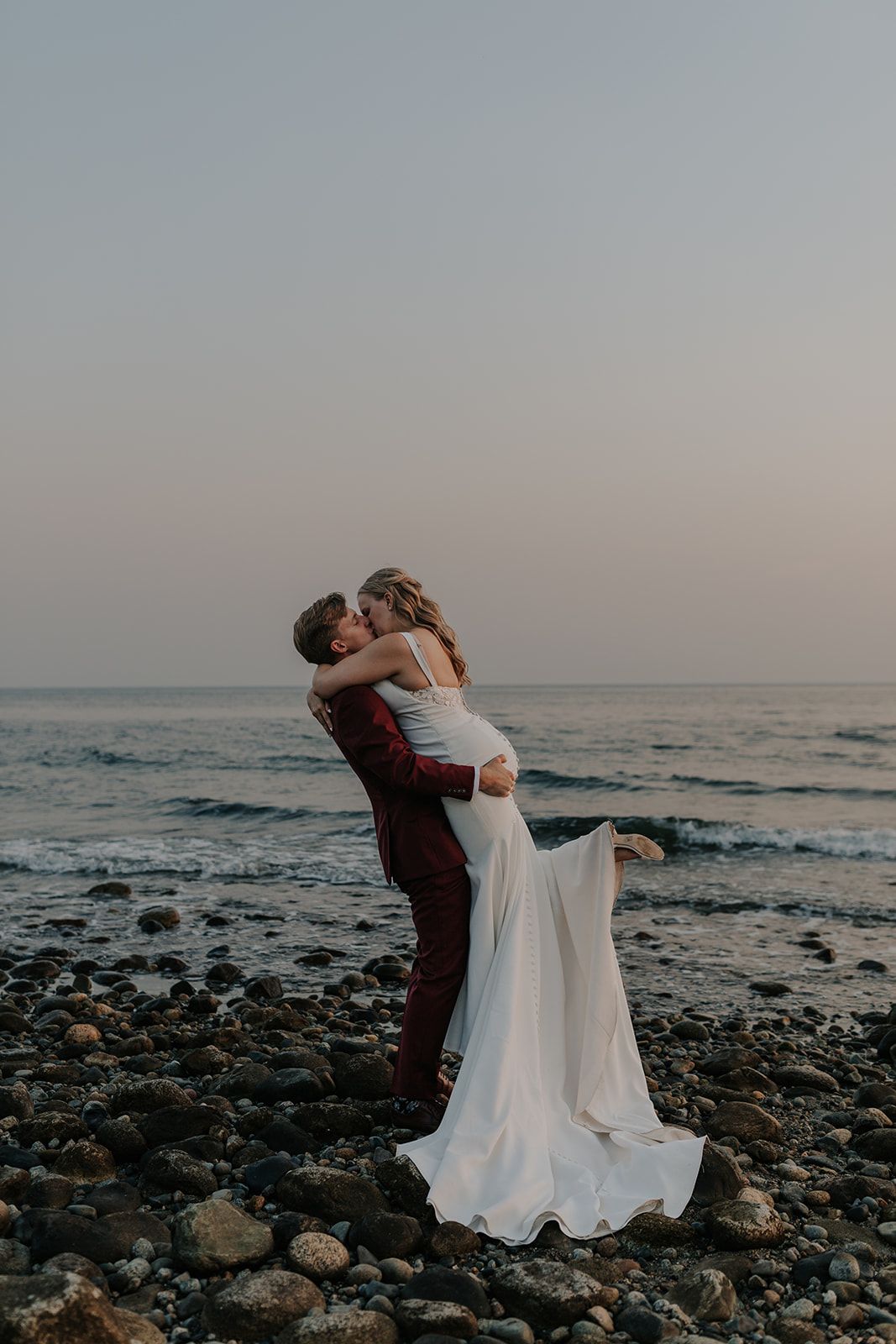 Couple in wedding attire embraces on a rocky beach at sunset.