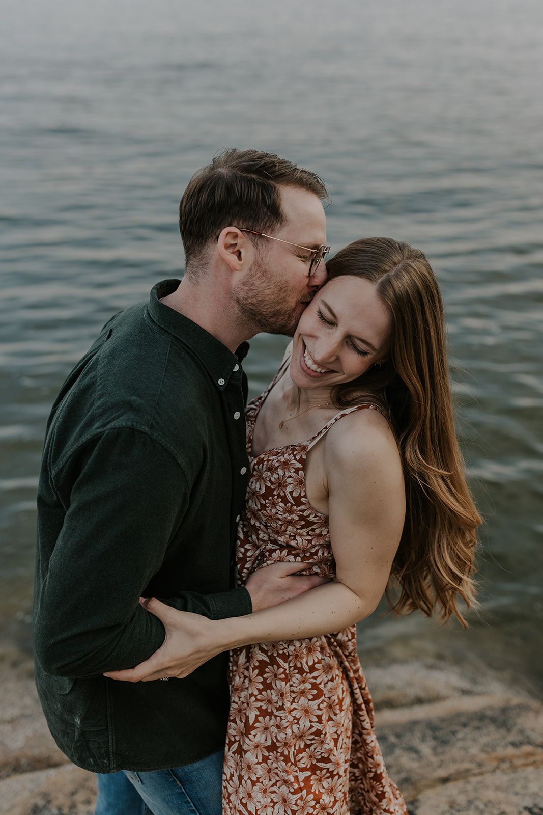 A couple shares a tender moment by the water, the man kissing the woman's temple as she smiles warmly in a floral dress.