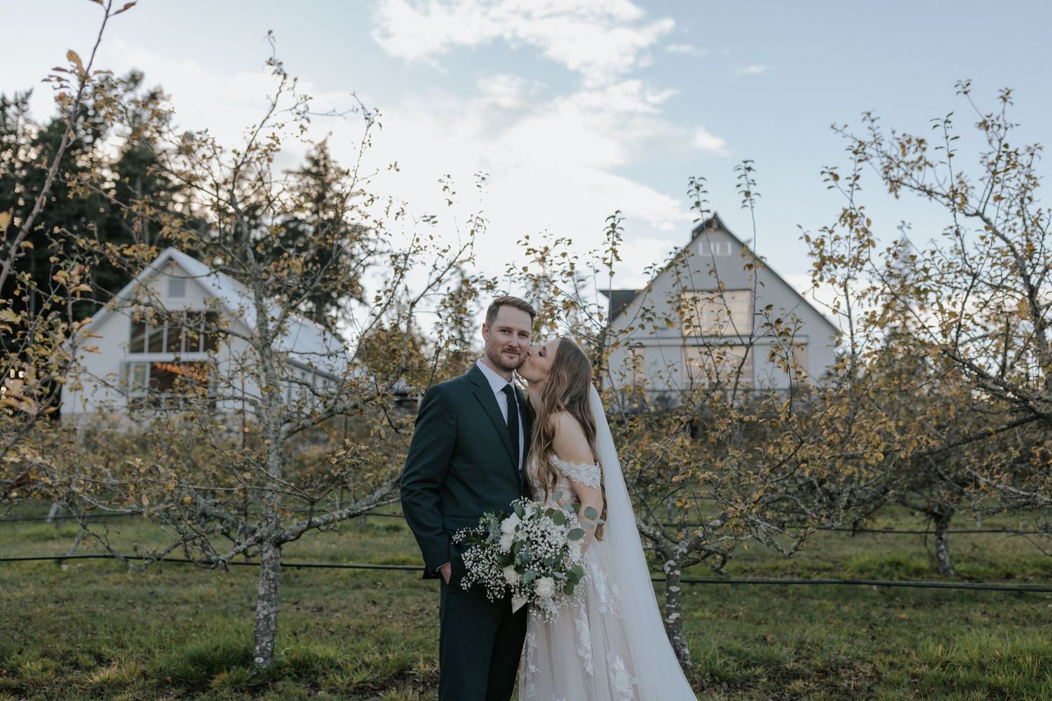 A couple embraces in a scenic apple orchard during sunset with white farmhouse buildings in the background.