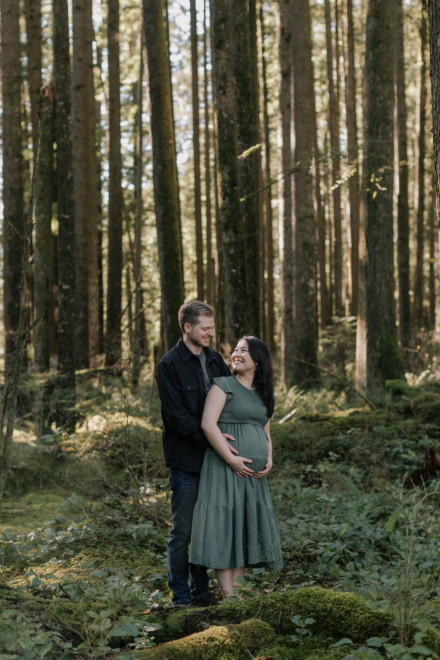 Couple poses in sunlit forest, pregnant woman in sage green dress, partner in black jacket embracing her from behind.