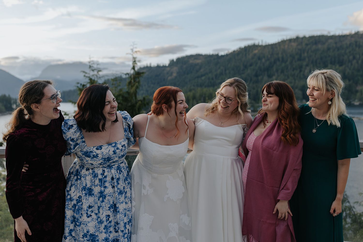 A group in mixed formal attire stands together on a deck with mountain views.