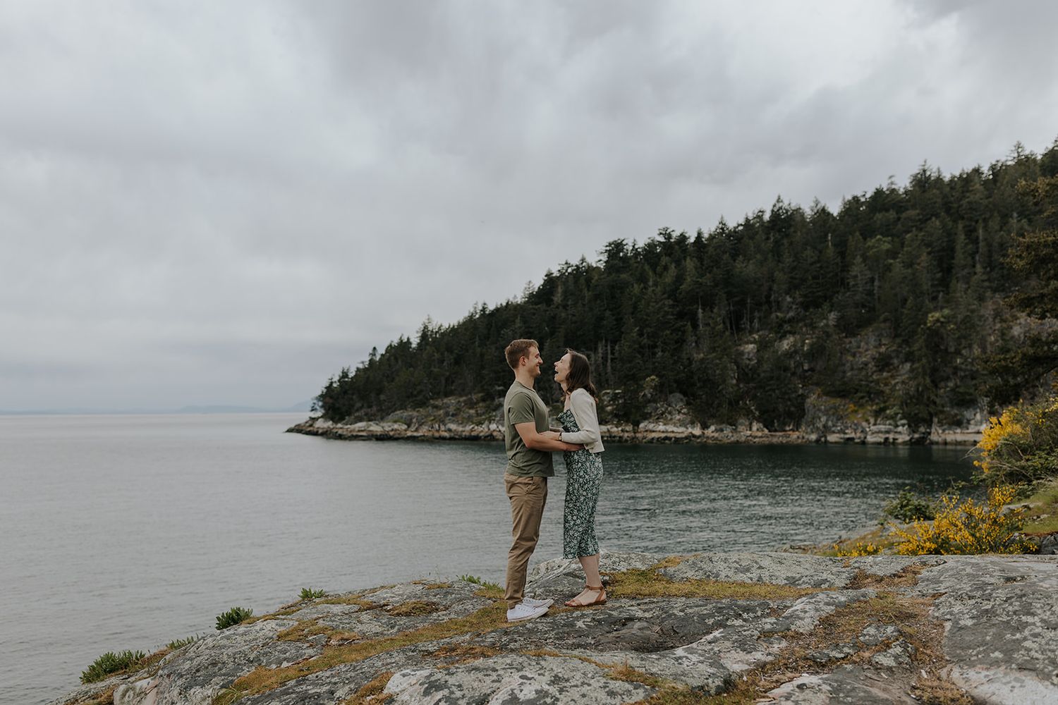A couple stands on rocky coastal cliffs overlooking a scenic ocean view.