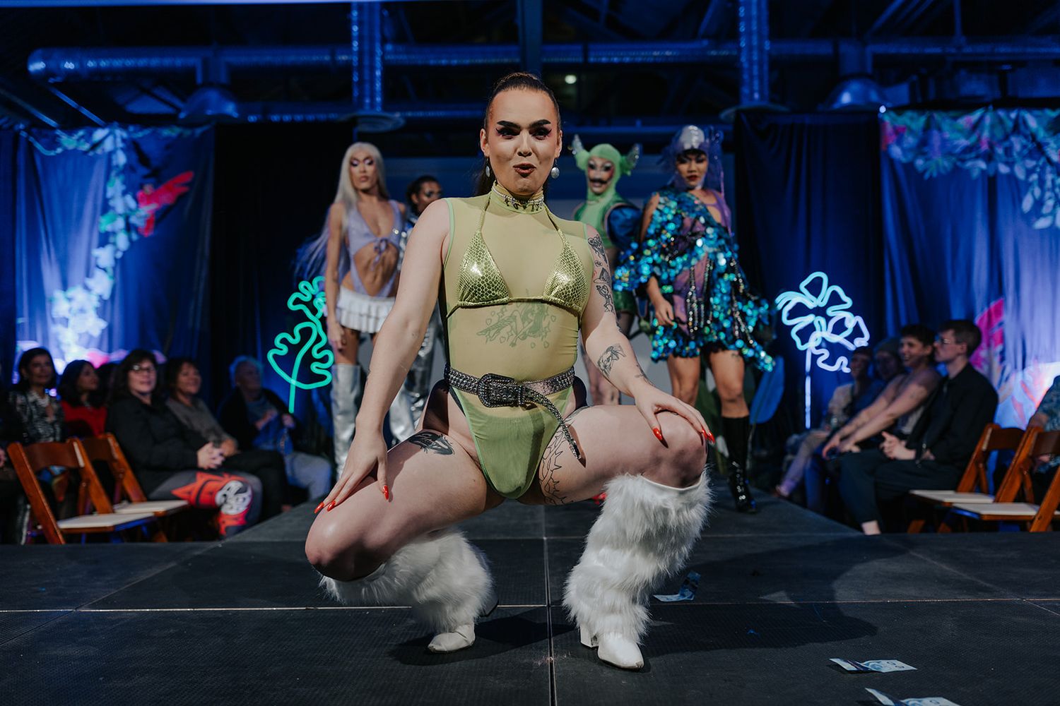 A performer in olive green outfit poses dramatically on stage with decorative lighting.