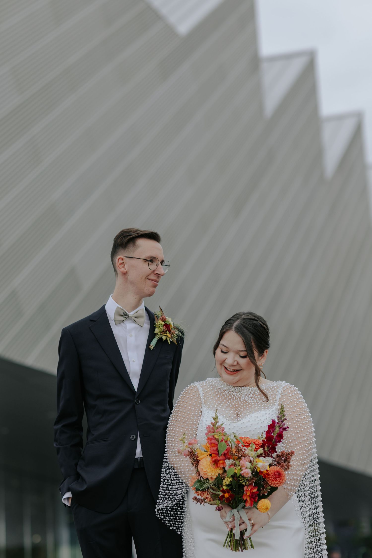 Wedding couple poses against a modern grey wall with the bride holding a bright fall-colored bouquet.
