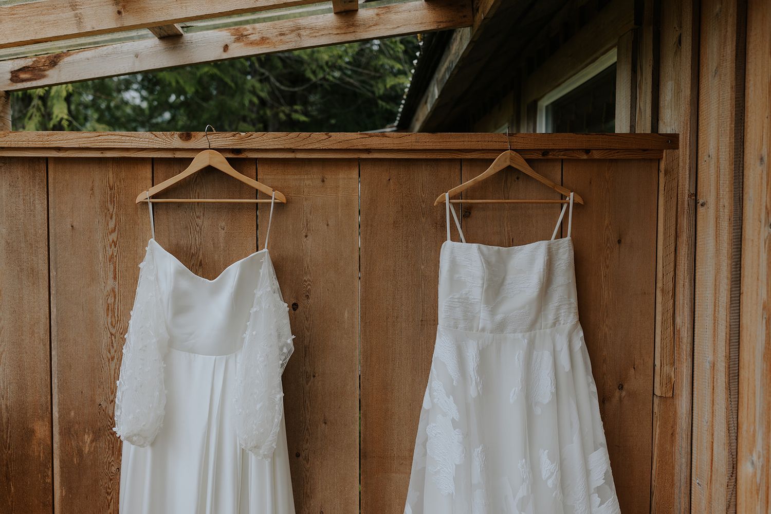 A row of white wedding dresses hanging on wooden hangers against a rustic wooden fence backdrop.