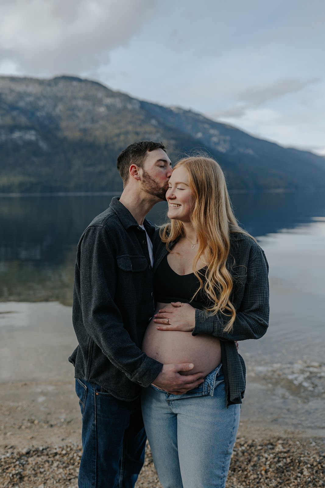 Intimate maternity photo of couple by a lake with mountains in the background.