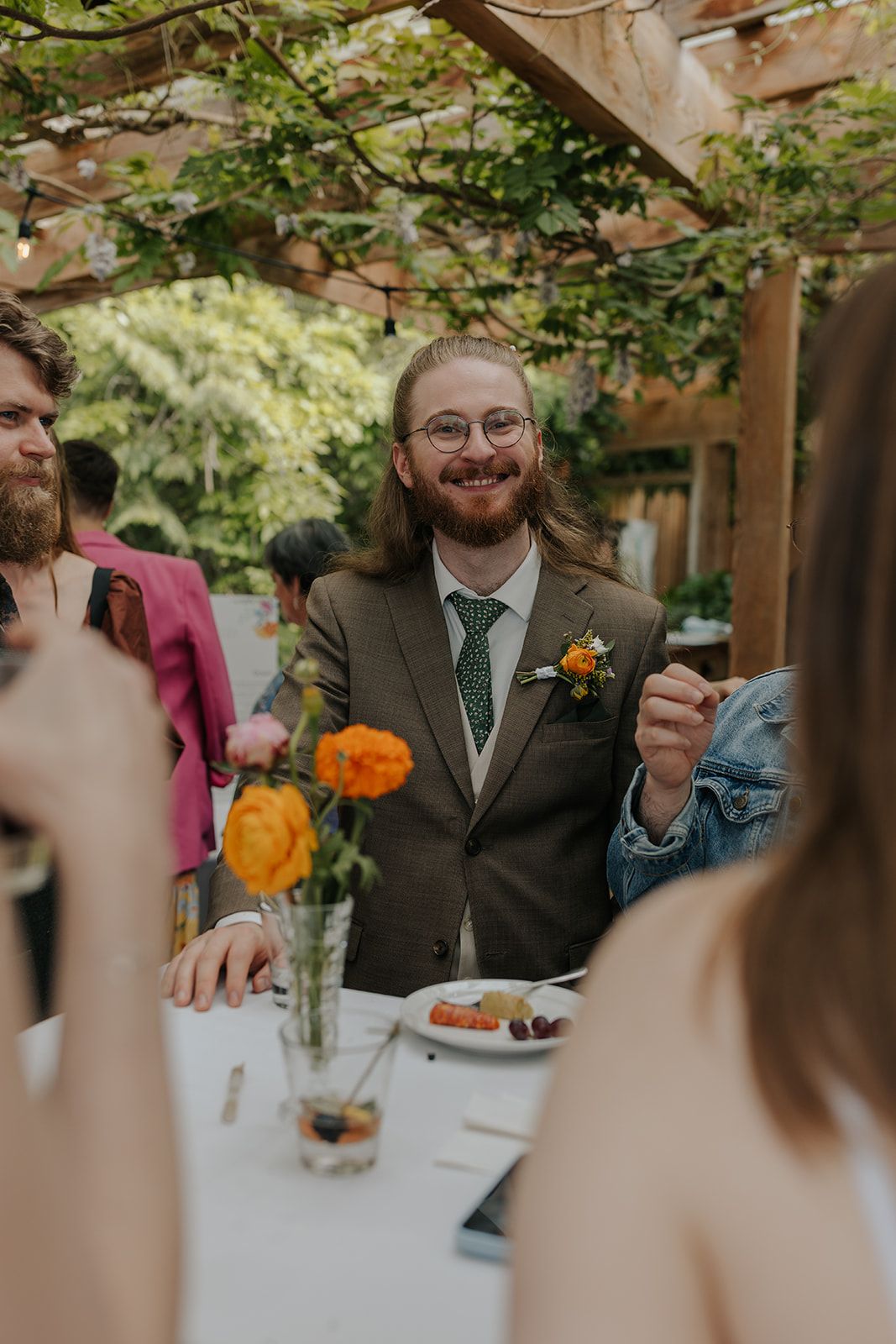 Guest in grey suit with orange boutonniere smiling during wedding reception.