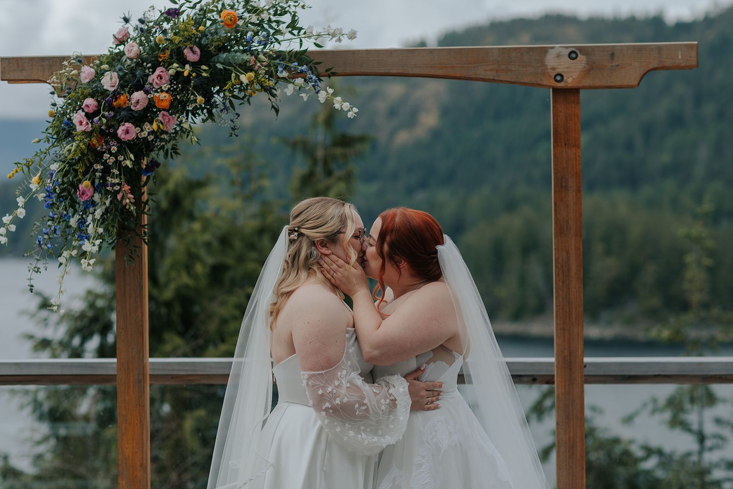 Wedding ceremony under wooden arch decorated with pink and orange flowers against lake view.