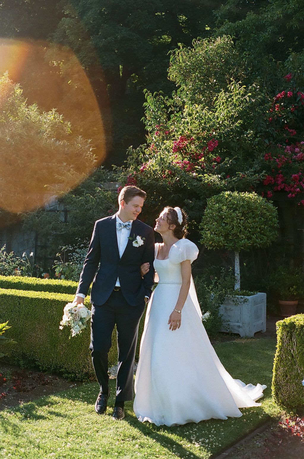 A couple walks through a sunlit garden during their wedding celebration.