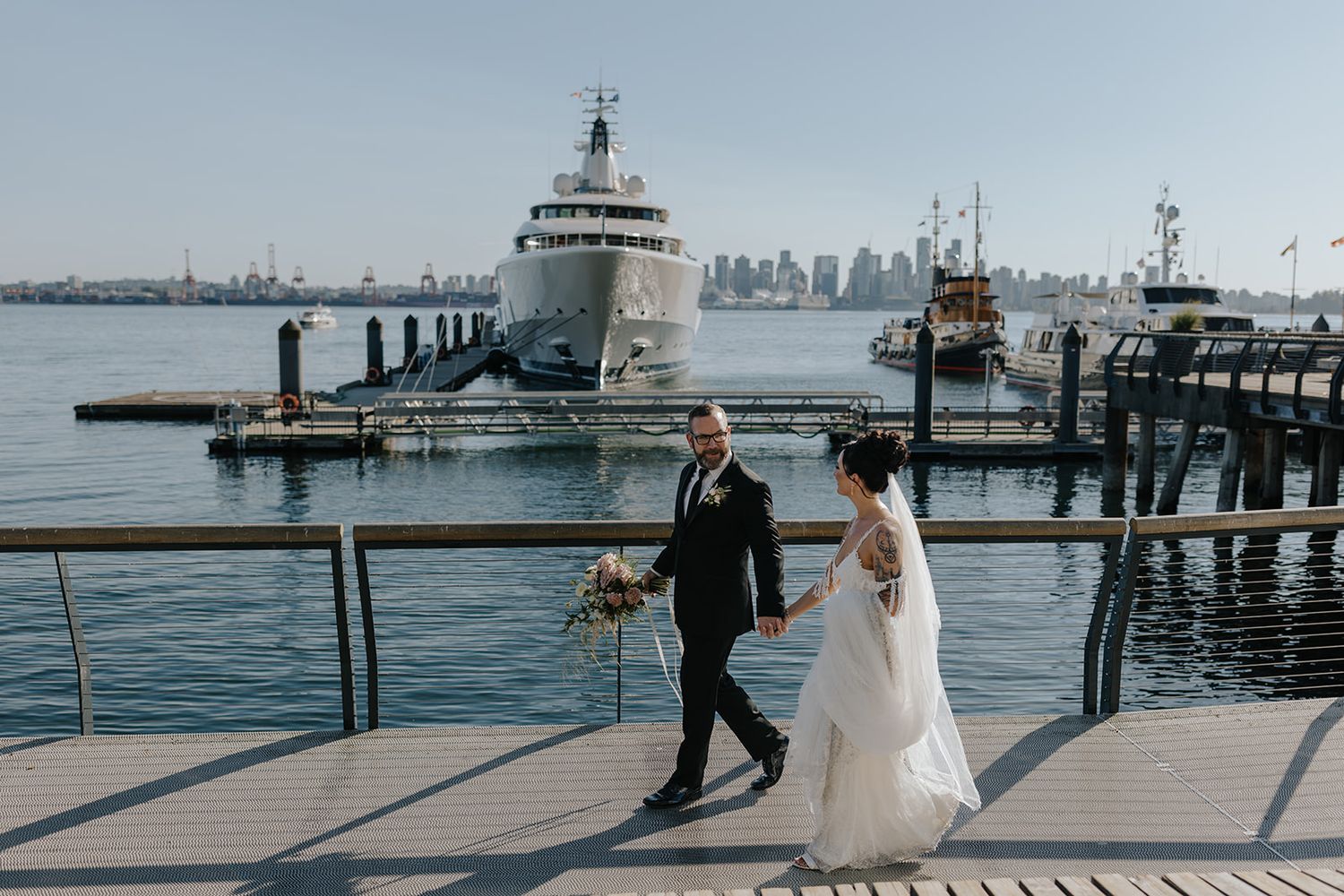 A couple in wedding attire walks along a waterfront pier with the city skyline visible in the distance.