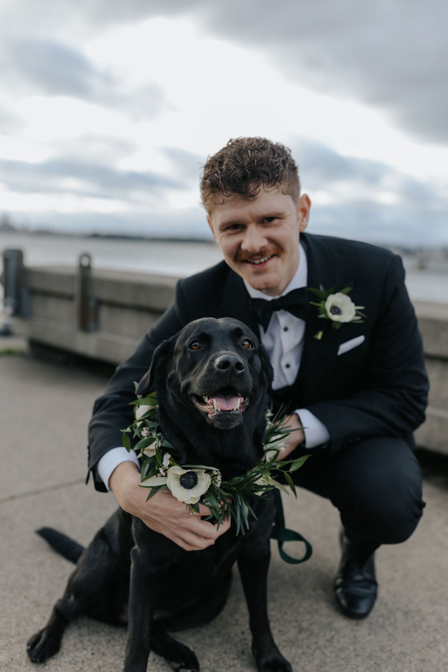 Black dog wearing floral collar poses with person in formal attire during outdoor wedding photoshoot.