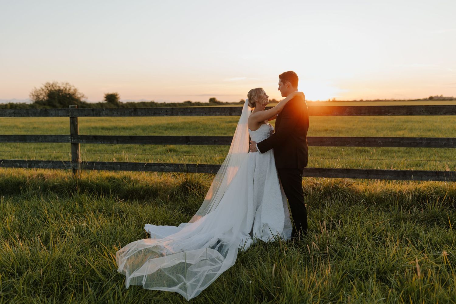 A couple in wedding attire embraces in a rural field at sunset with a flowing white dress trailing behind them.