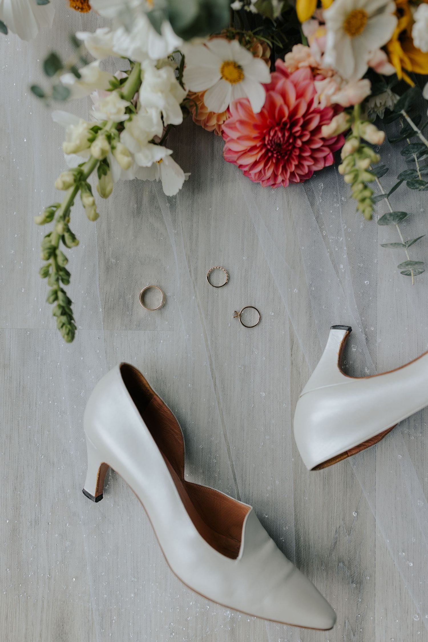 White bridal heels surrounded by colorful spring flowers and wedding rings on a light gray surface.
