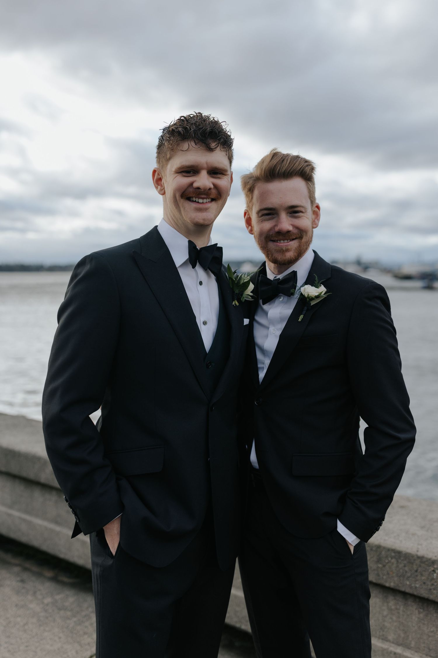 Groomsmen in black tuxedos share candid moments on cloudy waterfront.