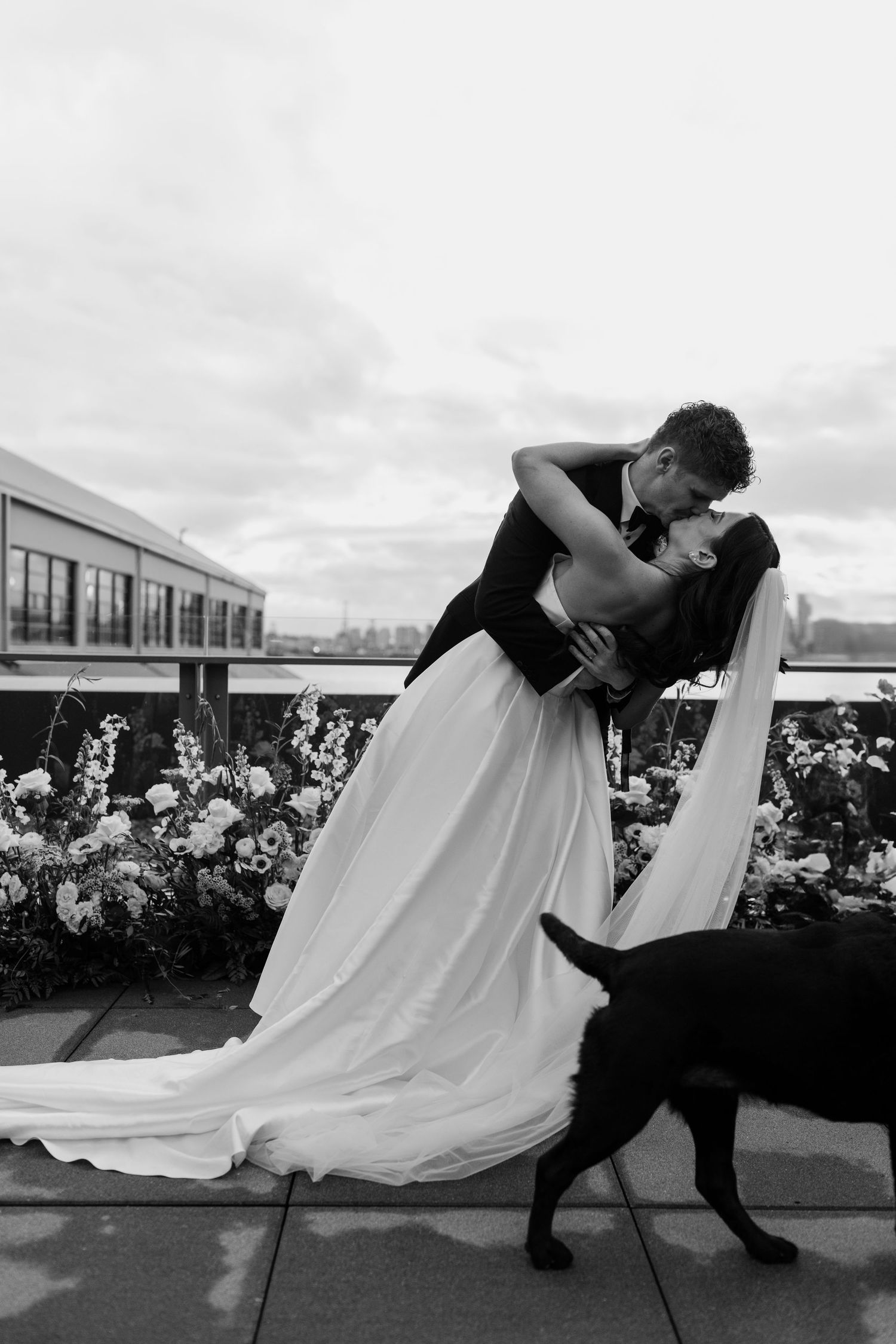Romantic black and white wedding photo with couple and their dog against urban backdrop.