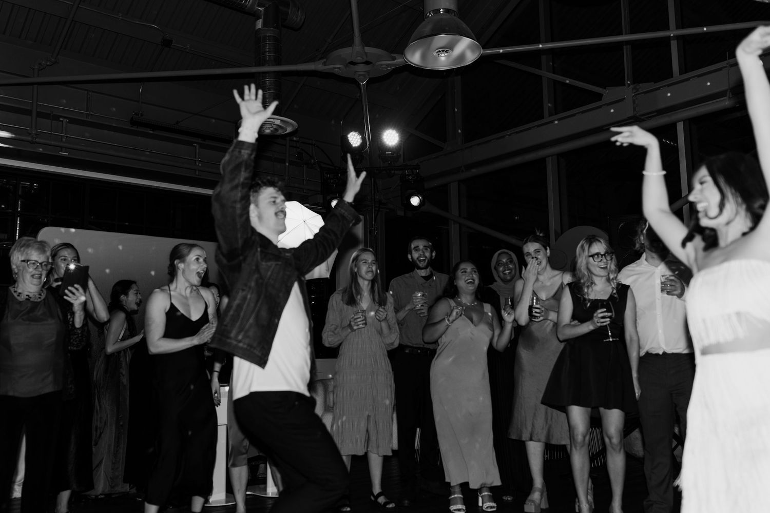 Wedding party and guests dance together in a dark reception venue with dramatic lighting and festive atmosphere.
