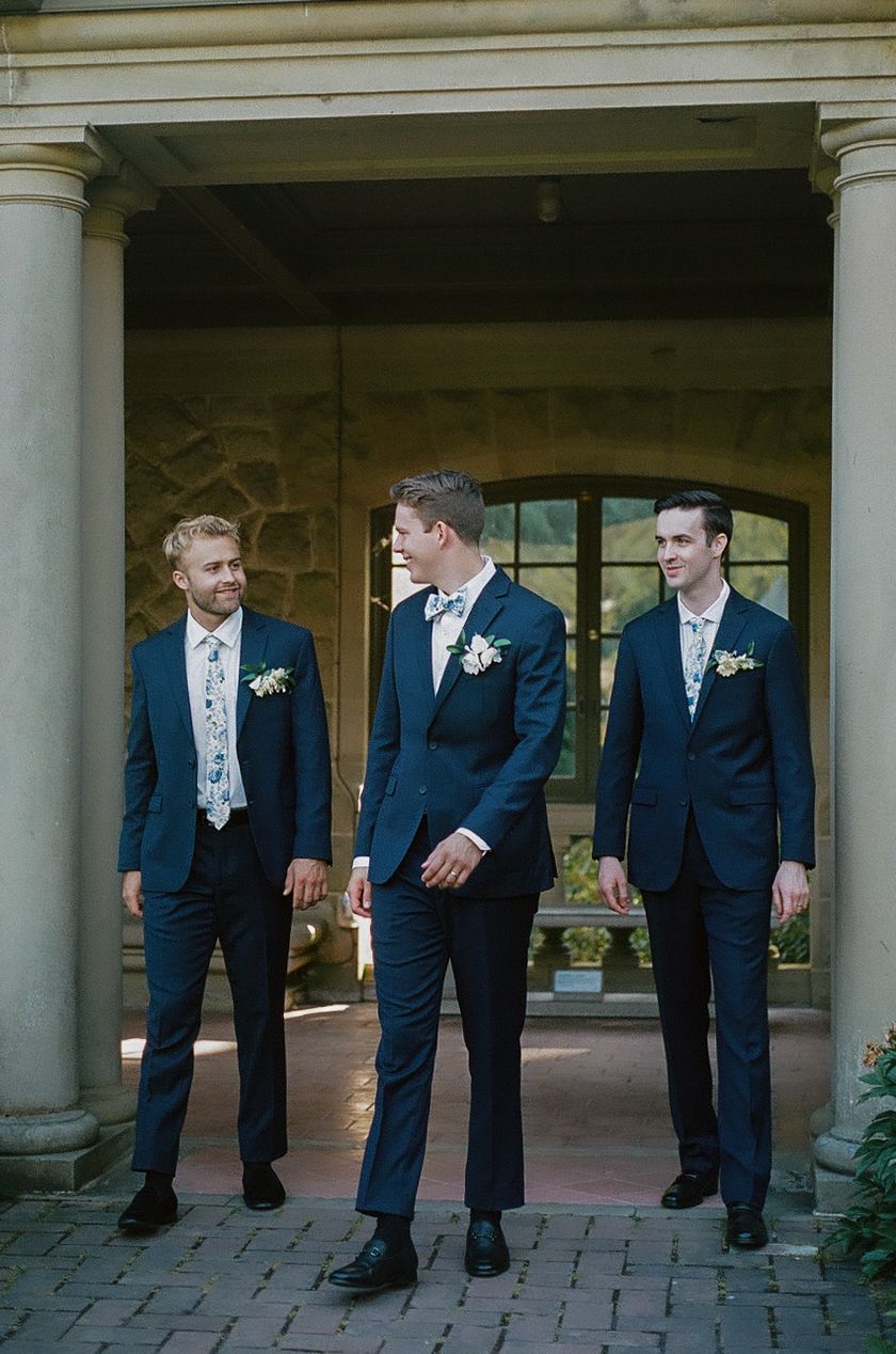 Three groomsmen in navy suits stand under a classical columned entrance.