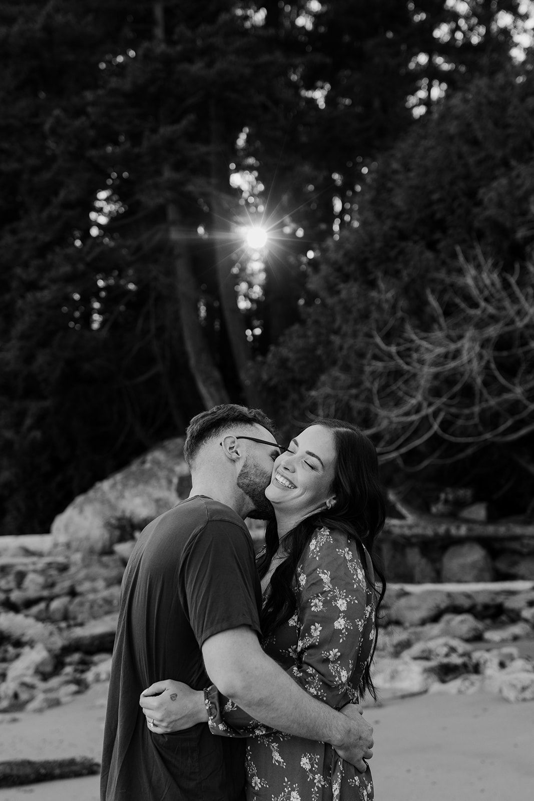 A couple shares joyful moments at sunset among trees in black and white photography.