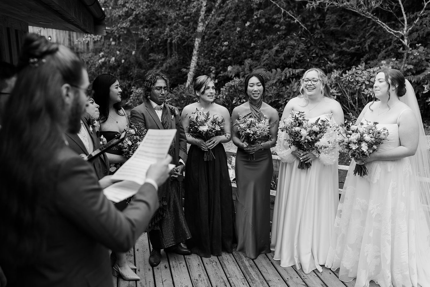 Wedding ceremony attendees standing in formal attire on wooden deck.