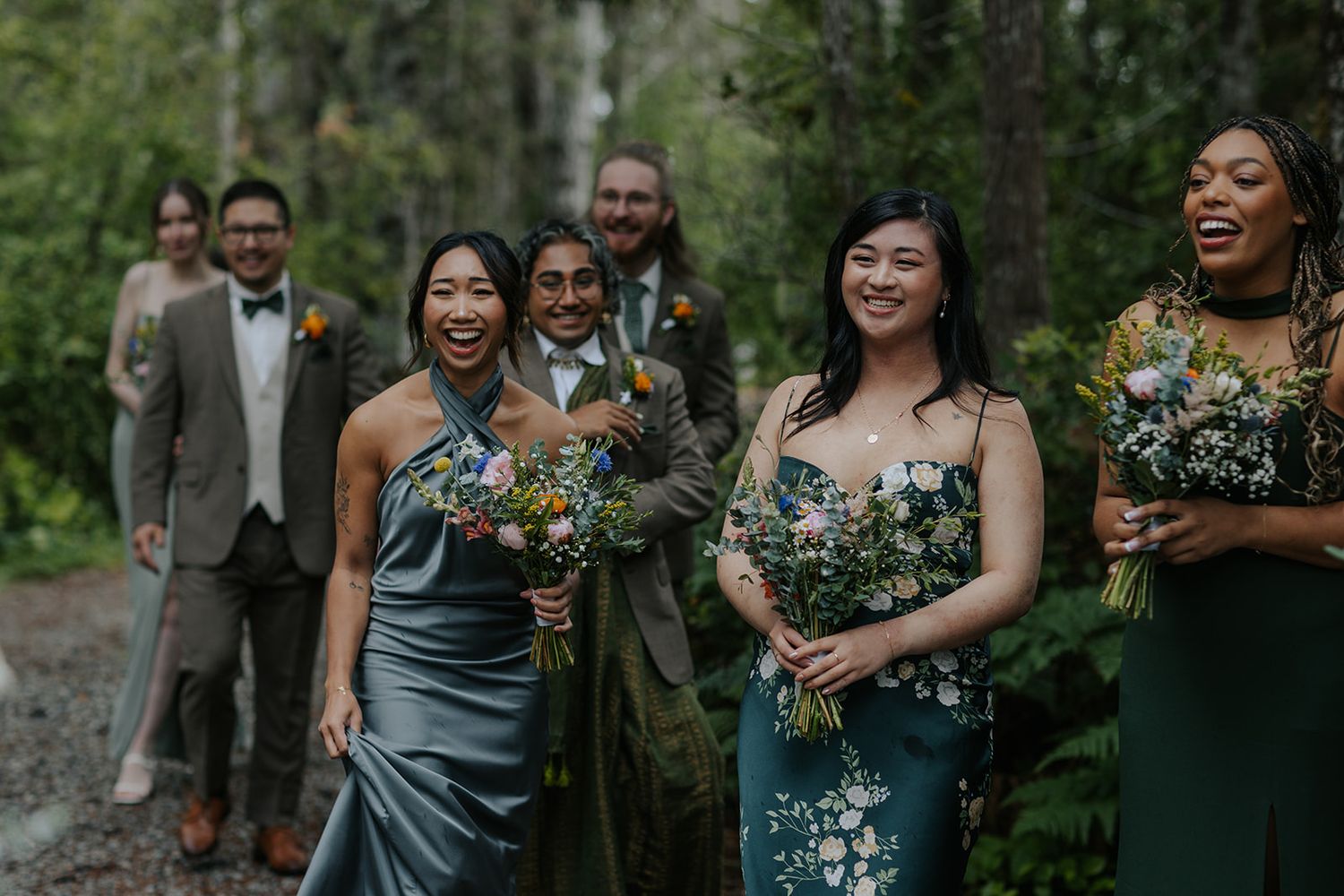 Wedding party laughing together in forest setting with mix of green and grey attire.