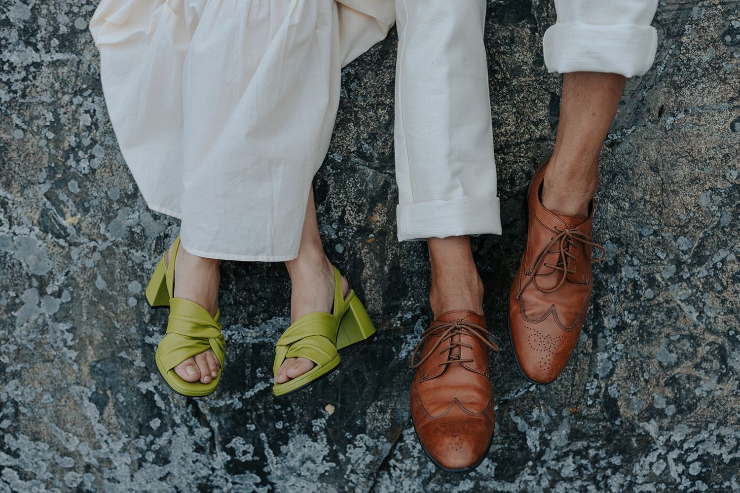 Wedding shoes detail shot showing yellow sandals and brown leather dress shoes on stone surface.