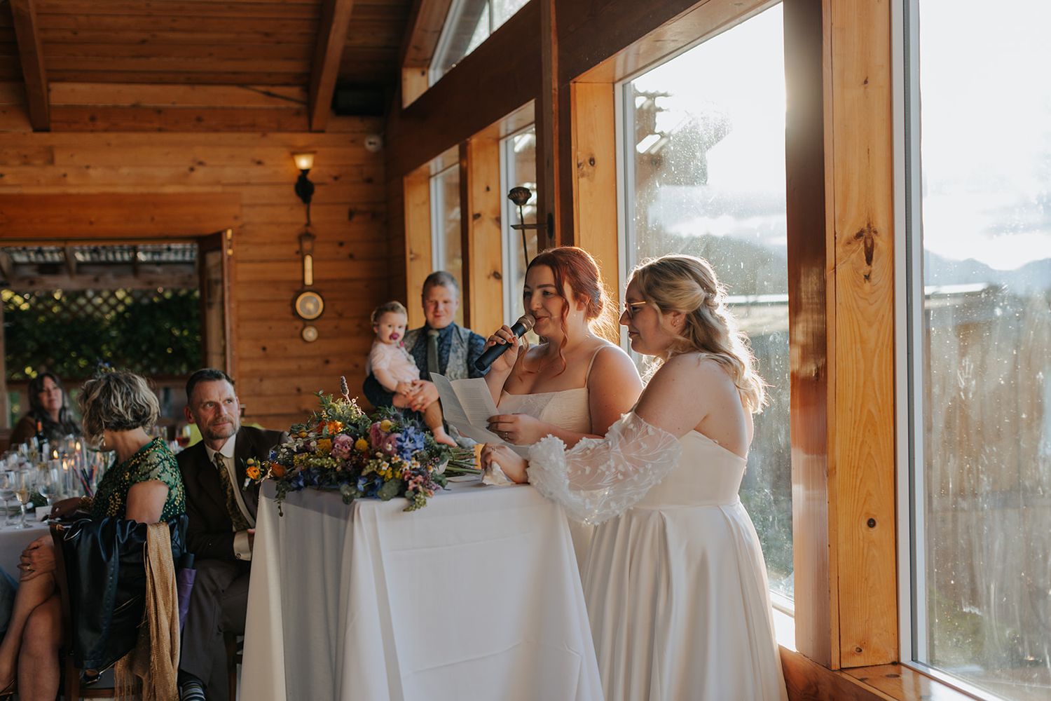 A wedding celebration in a rustic wooden venue with large windows overlooking mountains and water.