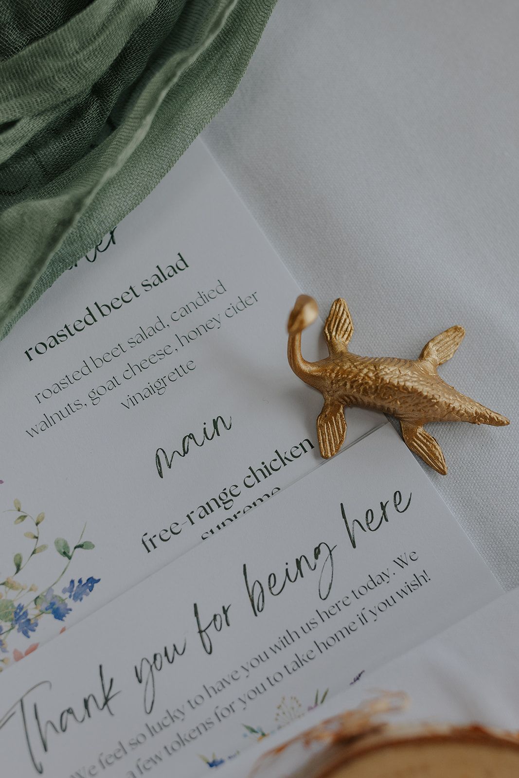 Close-up of a golden bird pin on a wedding menu card with green fabric accent.