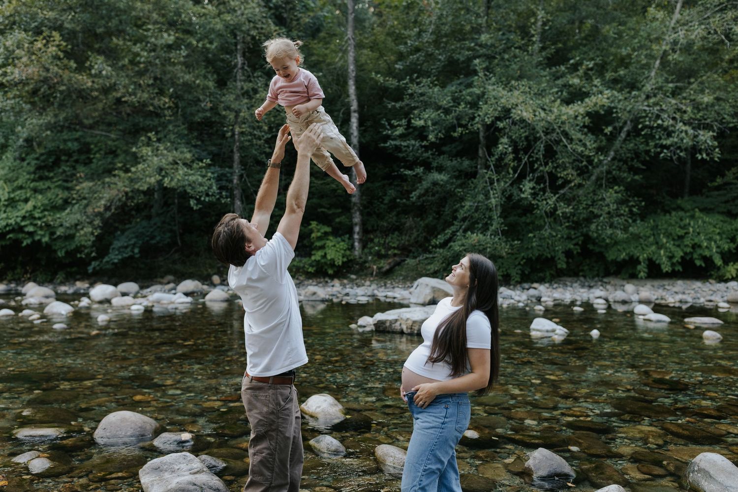 A father tosses his toddler in the air by a rocky river as his pregnant partner watches, surrounded by lush green trees.