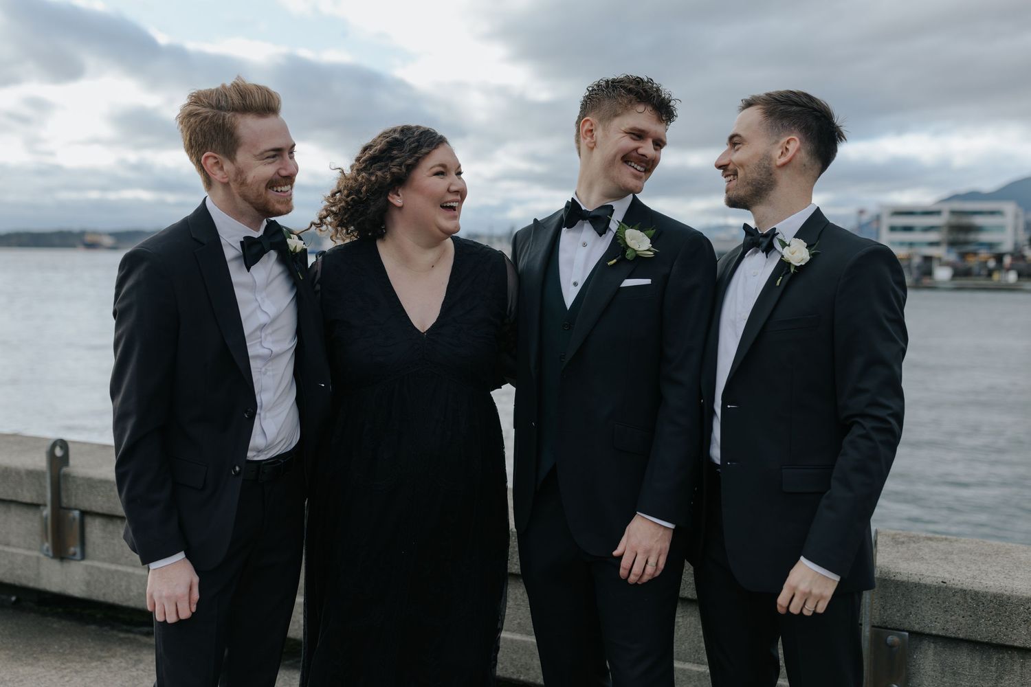 Groomsmen in black suits share laughs together on a cloudy day by the water.