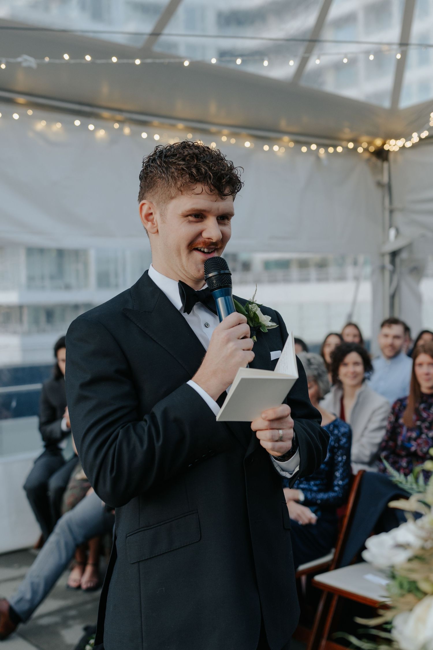 Wedding reception in tent with string lights as guest delivers emotional speech.