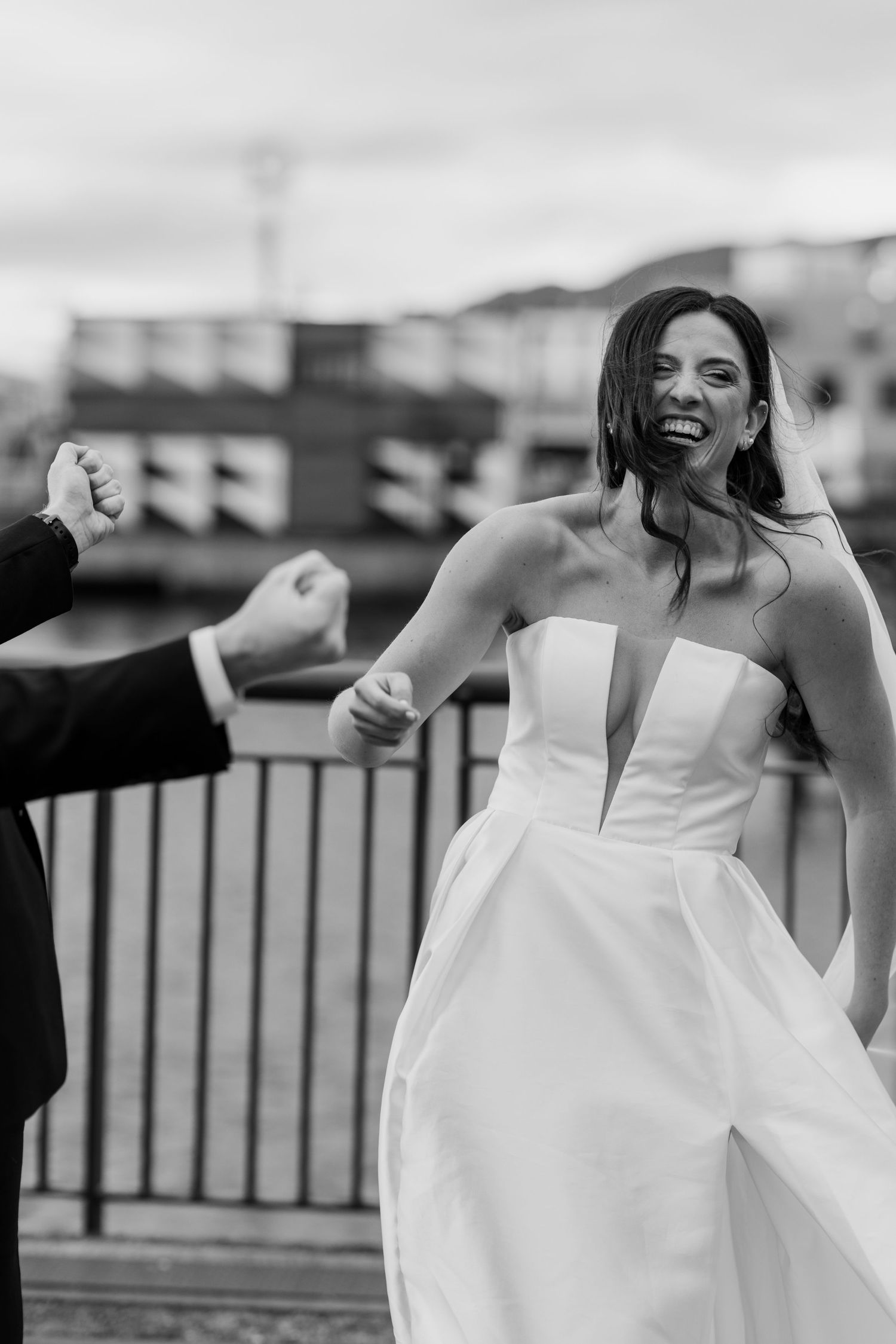 A bride in an elegant white dress laughs while dancing outdoors on a cloudy day.