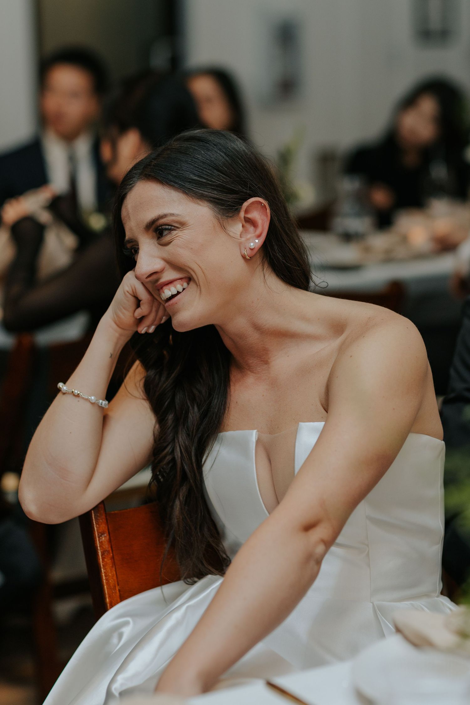A bride in a white strapless gown smiles joyfully while sitting during her wedding reception.