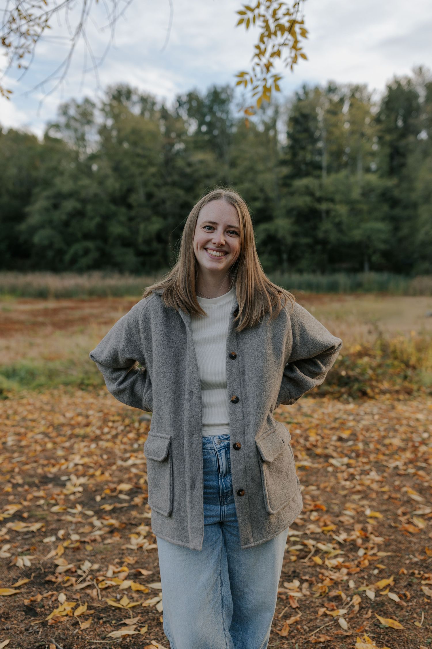 Smiling young woman in gray cardigan and jeans standing outdoors among autumn leaves with trees in background.