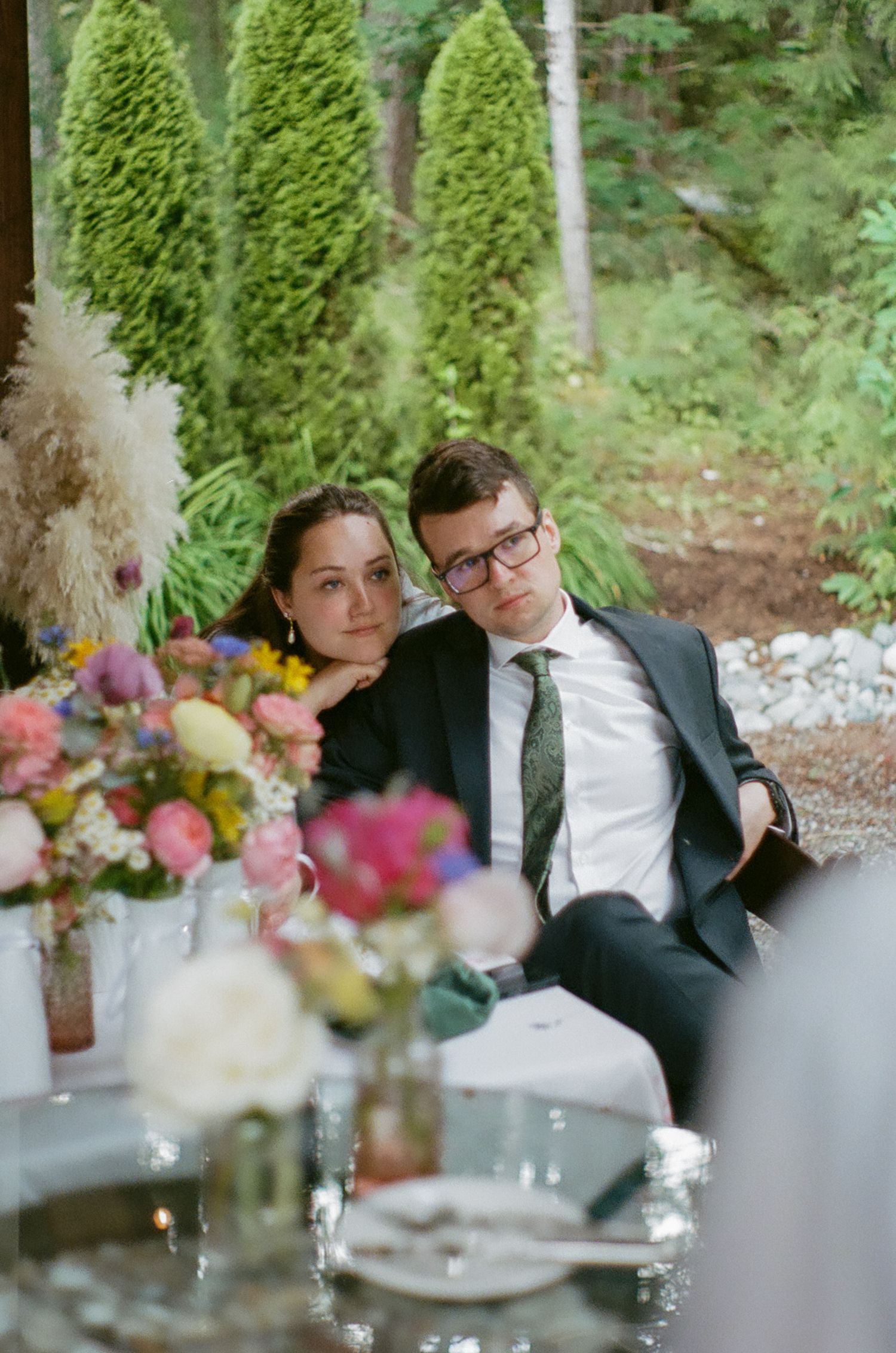 Wedding reception table setting with colorful floral centerpieces and evergreen trees in background.