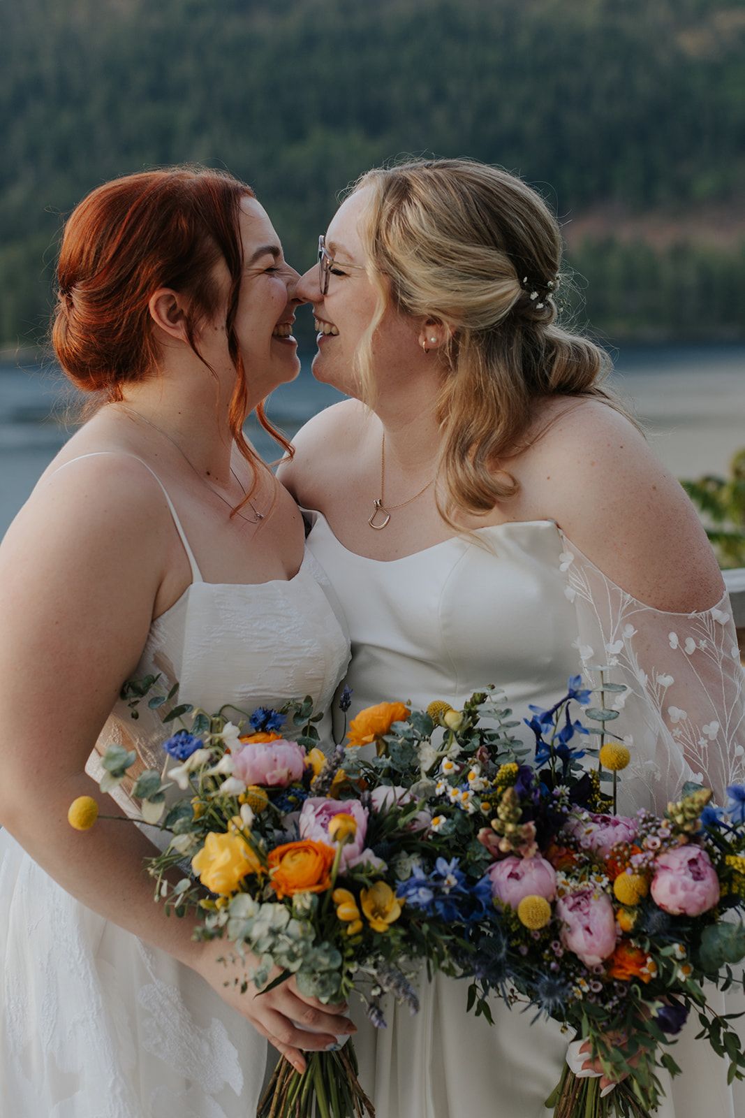 Wedding party members in white dresses holding vibrant multicolored bouquets against mountain scenery.