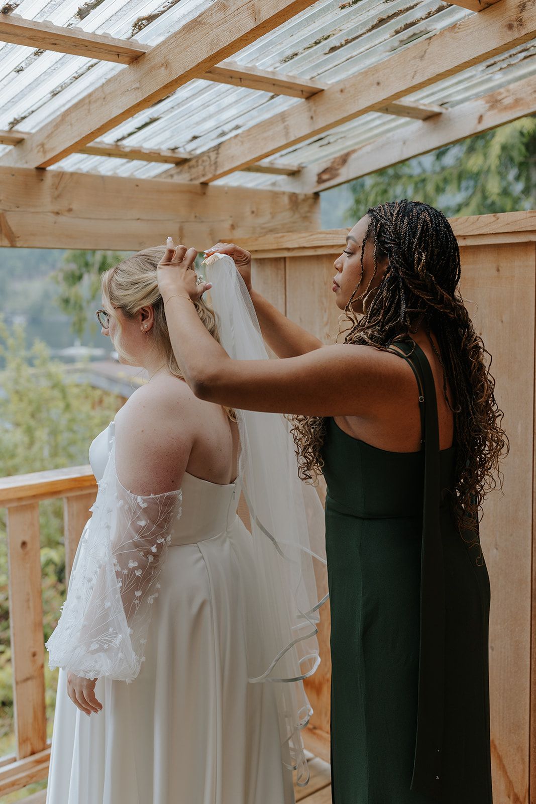 Helper in dark green dress adjusts wedding gown details on covered outdoor deck with mountain views.