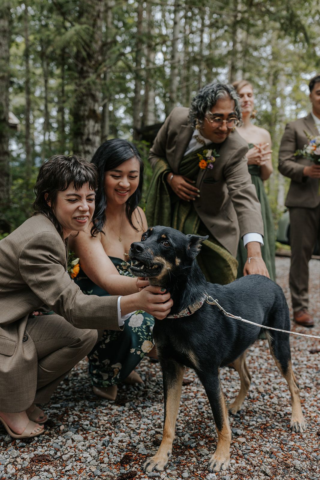 Wedding party members posing with a black dog on forest ground.