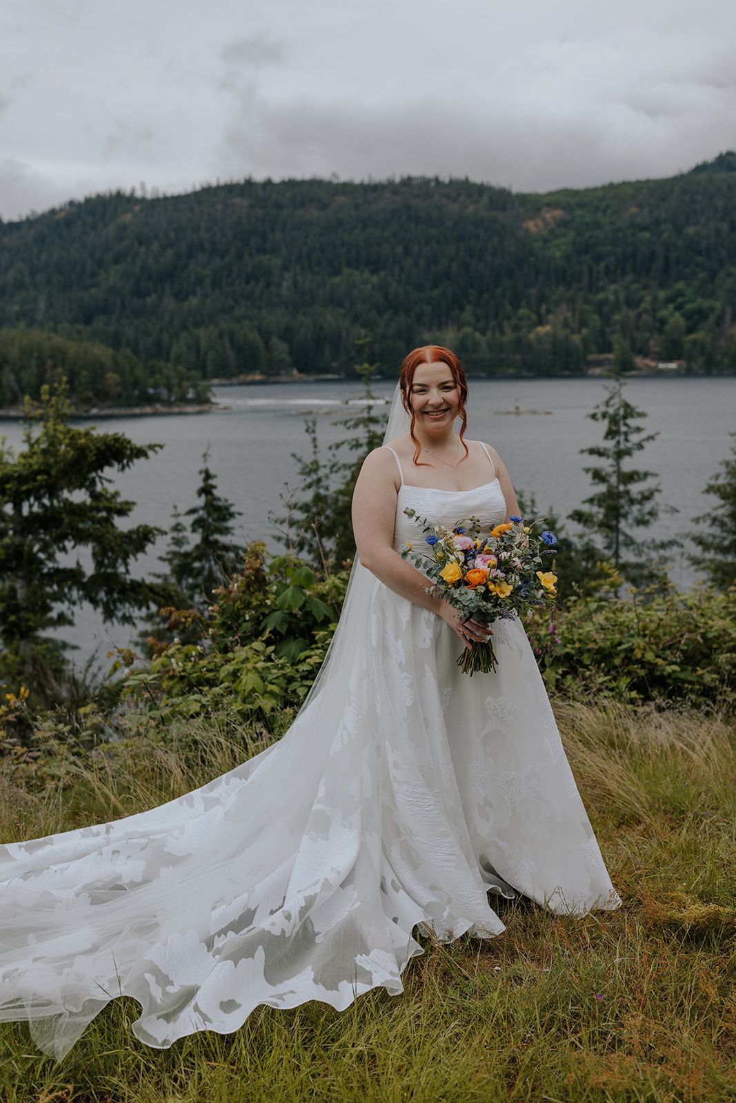 A person in a flowing white wedding dress holds a colorful bouquet against a mountain lake backdrop.